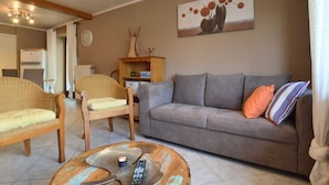 Una sala de estar con un sofá gris, dos sillones de mimbre, una mesa de centro con un control remoto y una vela, y un cuadro en la pared.