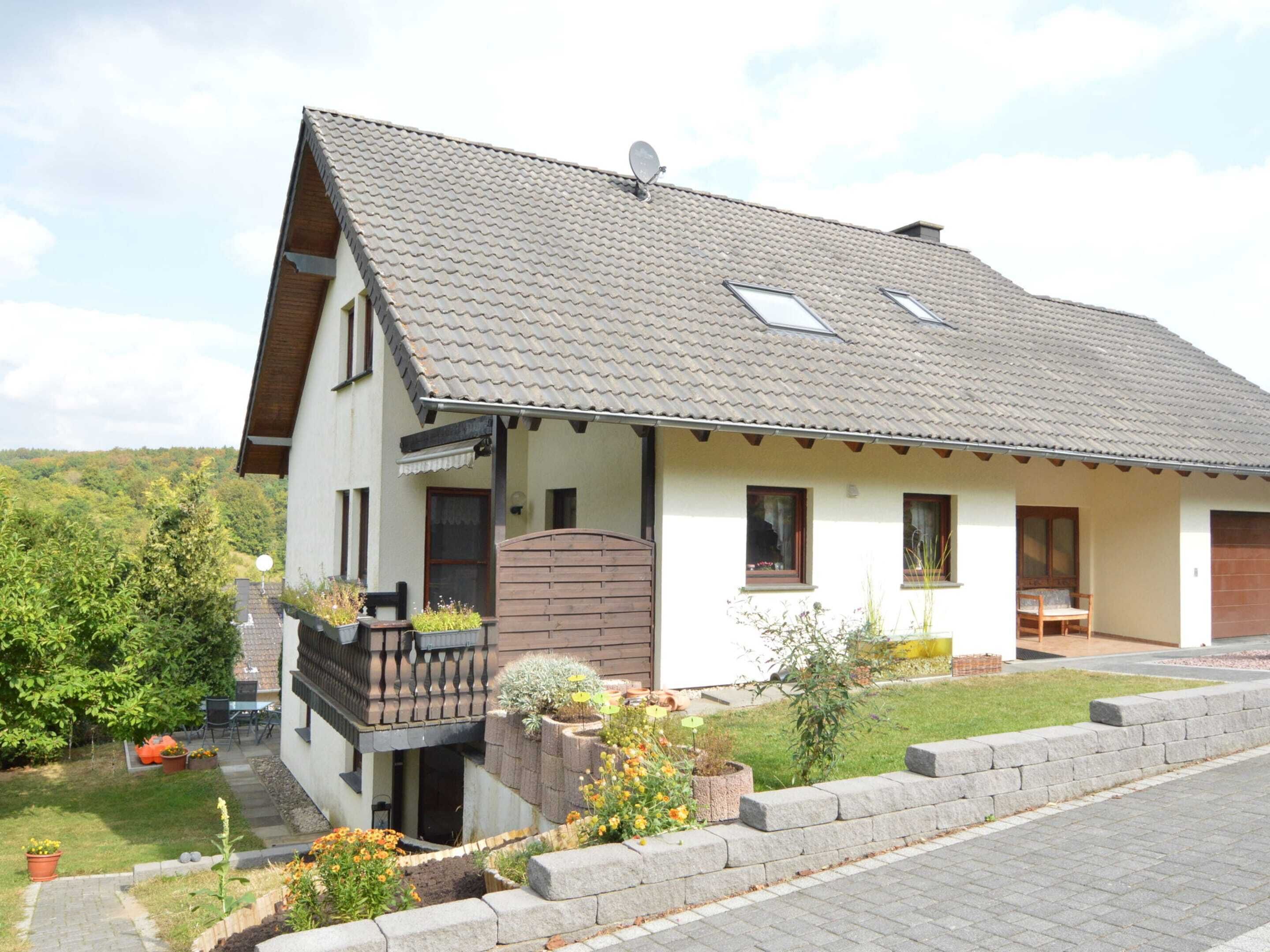 A two-story house with a sloped roof, a balcony, and a garden with various plants and flowers.
