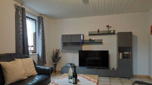 A living room with a leather sofa, a coffee table with a floral table runner, a flat-screen TV, and built-in shelving with decorative items. - Holiday Home Niederehe Romantic Escape (Üxheim)