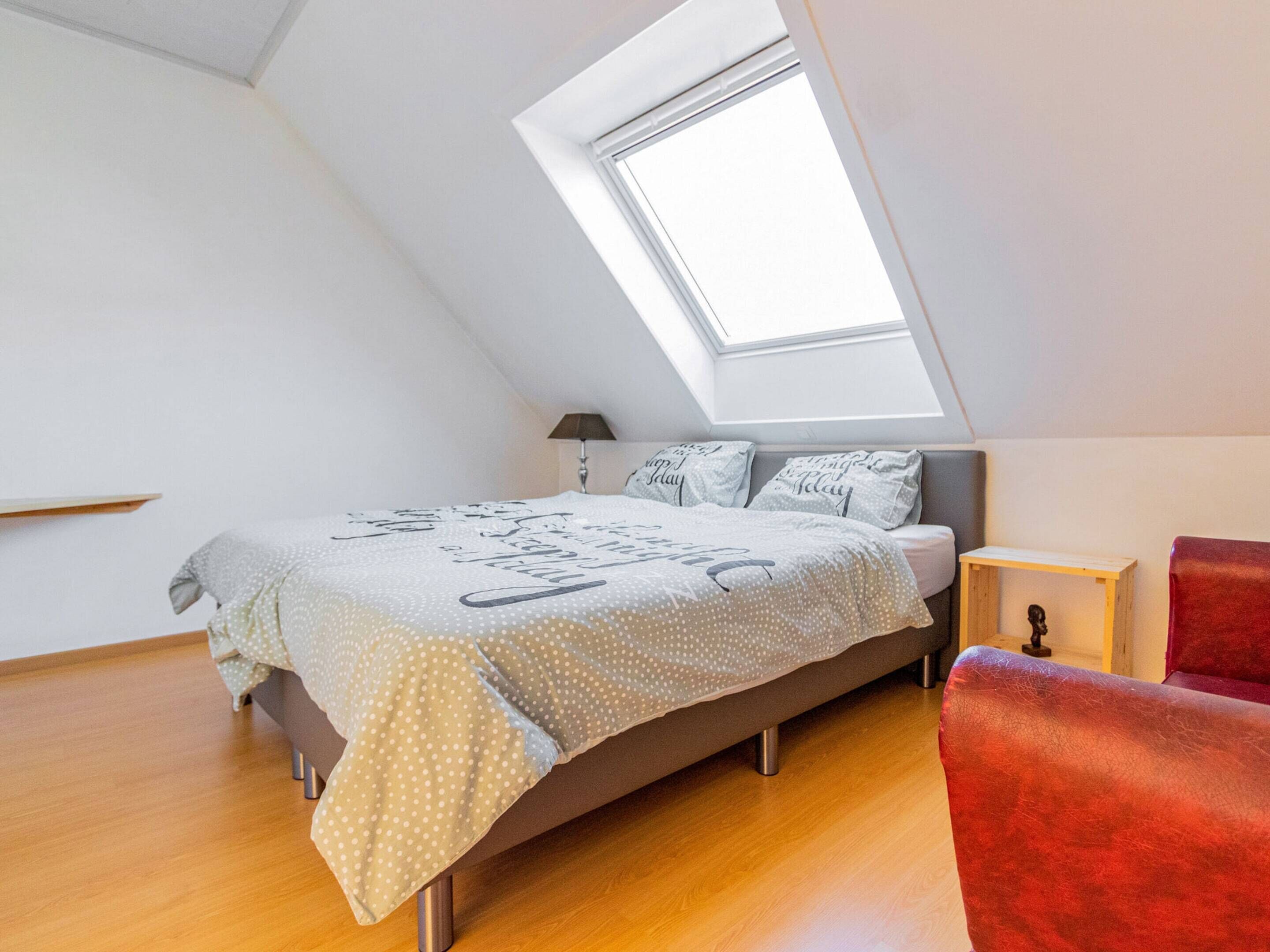A bedroom with a sloped ceiling, a skylight, a bed with a polka dot bedspread, a nightstand, and a red chair.