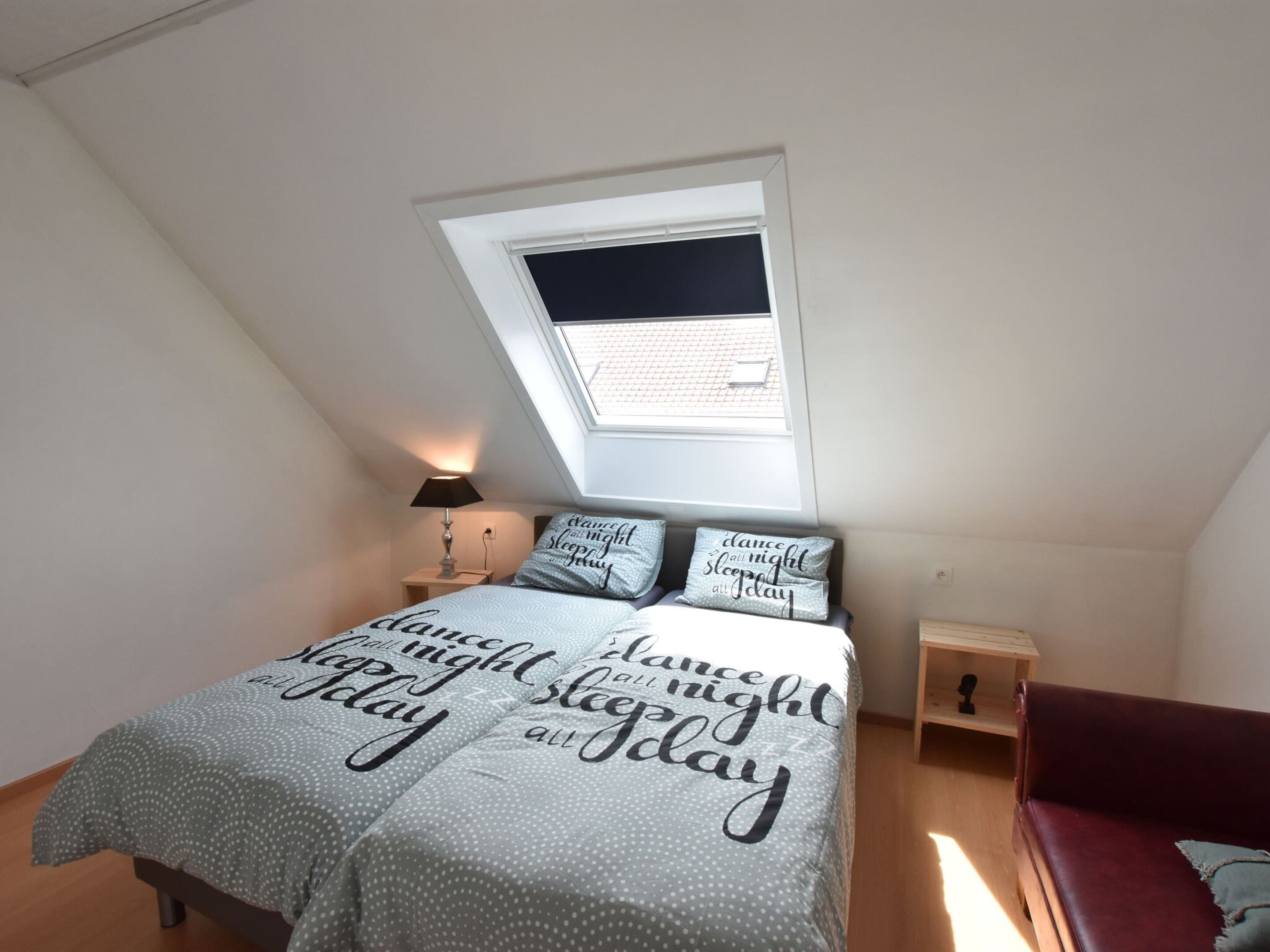 Ein Schlafzimmer mit einem Bett, einem Nachttisch, einer Lampe, einem Fenster und einem roten Sofa.