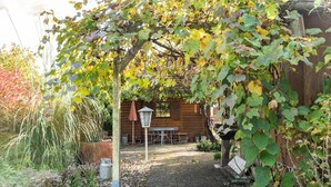 A garden with a pergola covered in vines, a wooden building, and a table with chairs in the background. - Charming Holiday Home With Private Garden (Niederaula)