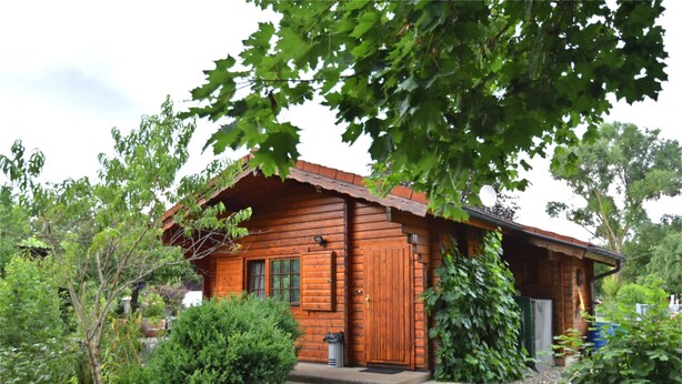 A wooden cabin with a sloped roof, surrounded by dense greenery and a gravel driveway. - Charming Holiday Home With Private Garden (Niederaula)