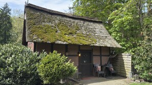 Um edifício rústico com telhado de palha coberto de musgo, fachada de tijolo e uma pequena área externa com cadeiras e mesas.