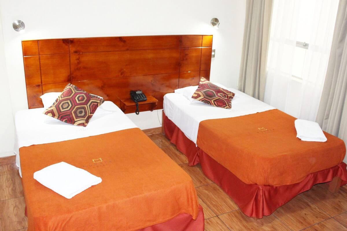 Habitación de hotel con dos camas individuales, cada una con una colcha naranja y almohadas a juego. Se aprecia un cabecero de madera, una mesita de noche con un teléfono y una ventana con cortinas.