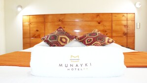 A hotel bed with a wooden headboard, two patterned pillows, and a white bedspread with the hotel name 'MUNAYKI HOTEL' on it. - Munayki Hotel (Tacna)