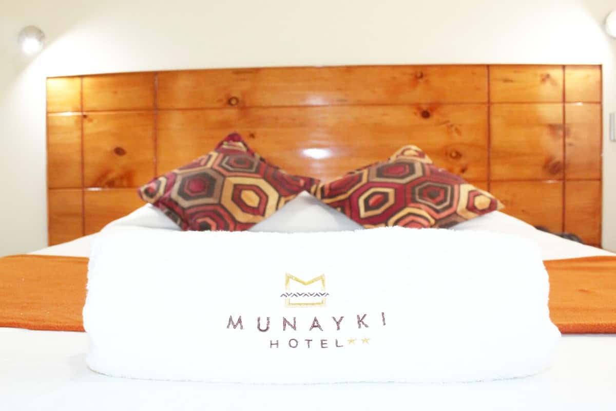 Una cama de hotel con cabecera de madera, dos almohadas estampadas y una colcha blanca con el nombre del hotel 'MUNAYKI HOTEL'.