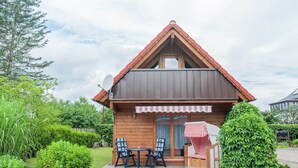 Une cabane en bois avec un toit en pente, une véranda avec deux chaises et une table, et un petit jardin agrémenté d’une haie et d’un arbre.