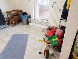 A room with a tiled floor, a blue rug, a red tricycle, a wooden cart, a trash bin, and a radiator. - Flat Near the ski Area in Medebach (Medebach)