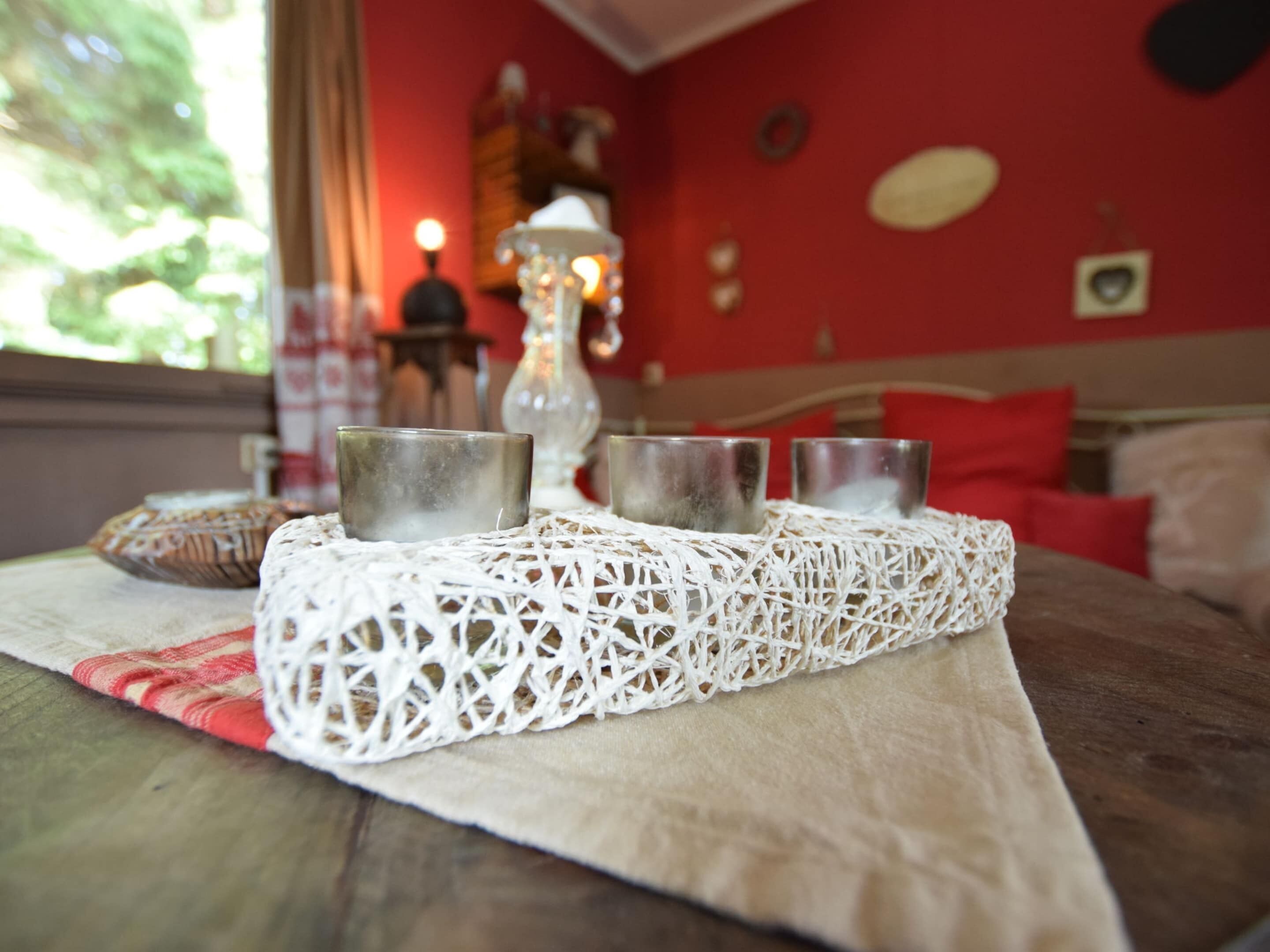 A table with a white lace table runner, three metal cups, and a loaf of bread on a red tablecloth.