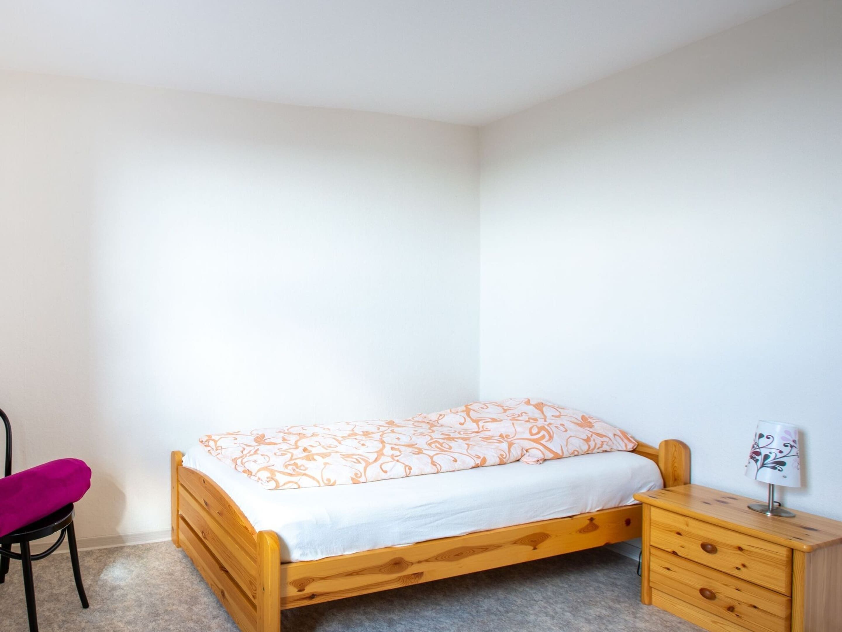 A small, neatly arranged bedroom with a single bed, a wooden nightstand, a chair, and a lamp.