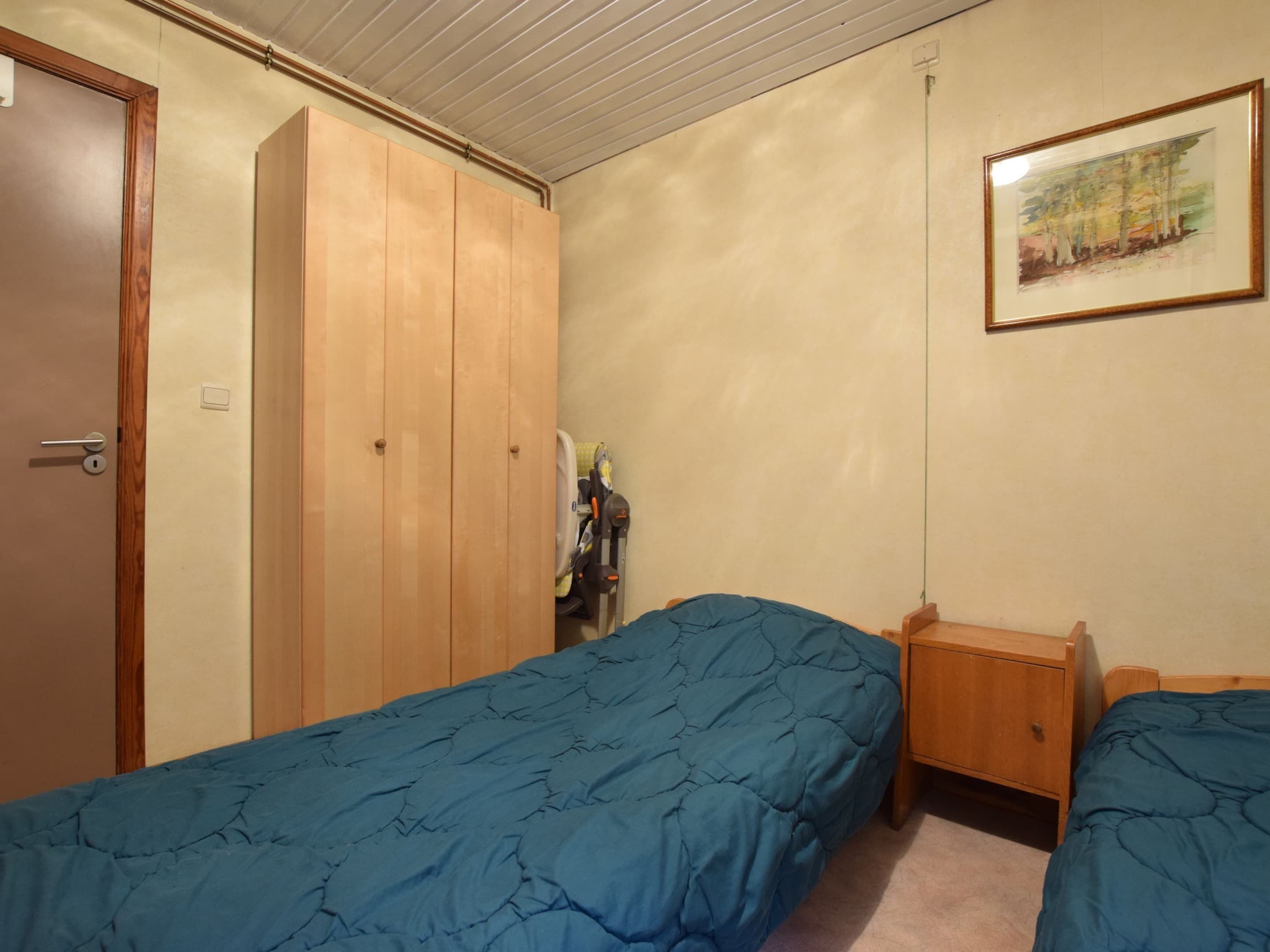 A bedroom with two single beds, a wooden wardrobe, a nightstand, and a framed painting on the wall.