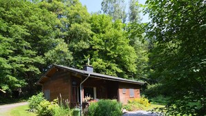 A rustic cabin with a chimney, surrounded by dense greenery and a gravel path leading to the entrance. - Forest Cottage With Ponds (Bouillon)