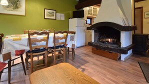 A dining area with a table set for four, wooden chairs, a fireplace, and a kitchen in the background. - Forest Cottage With Ponds (Bouillon)