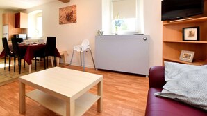 Un soggiorno con pavimento in legno, un'area pranzo con tavolo e sedie, un termosifone, una TV su uno scaffale e una finestra con una pianta.