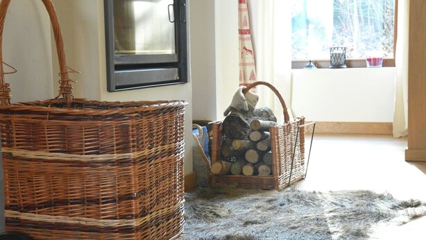 A room with a large wicker basket, a smaller wicker basket with logs, a fur rug, and a window with curtains.