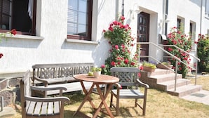Een terras met een houten bank en tafel, omgeven door een wit gebouw met een trap en bloembakken.