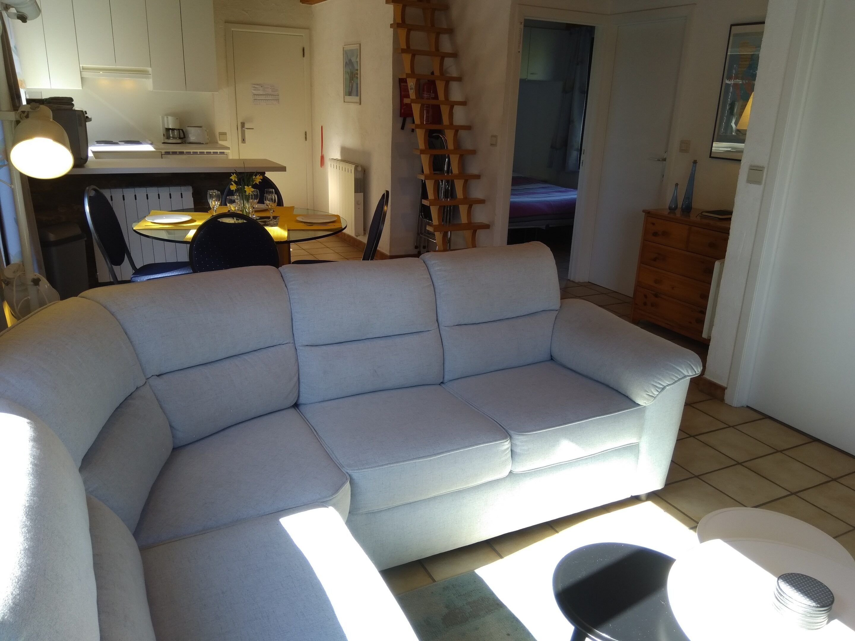 Una sala de estar con un sofá gris, una mesa de comedor con sillas, una zona de cocina con armarios y un radiador, y una escalera de madera.
