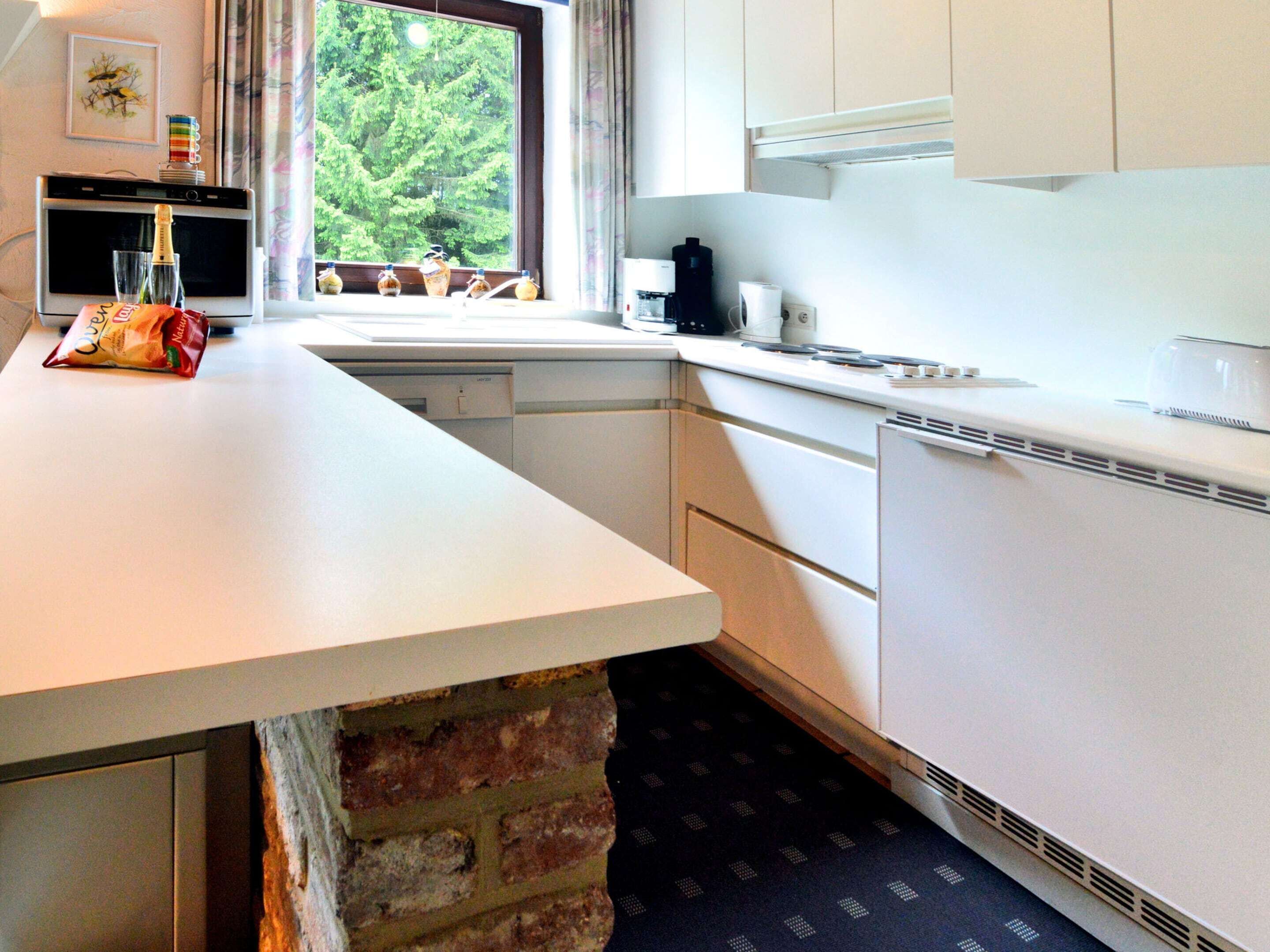 A kitchen with white cabinets, a brick wall, a window with a view of trees, and a countertop with a coffee maker and a bag of chips.