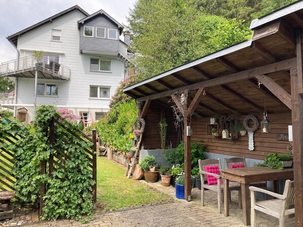 A wooden pergola with a table and chairs, surrounded by potted plants and a brick pathway.