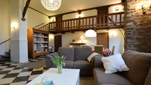 Un salon chaleureux avec un sol à carreaux, un escalier en bois, un lustre et un mur en pierre.