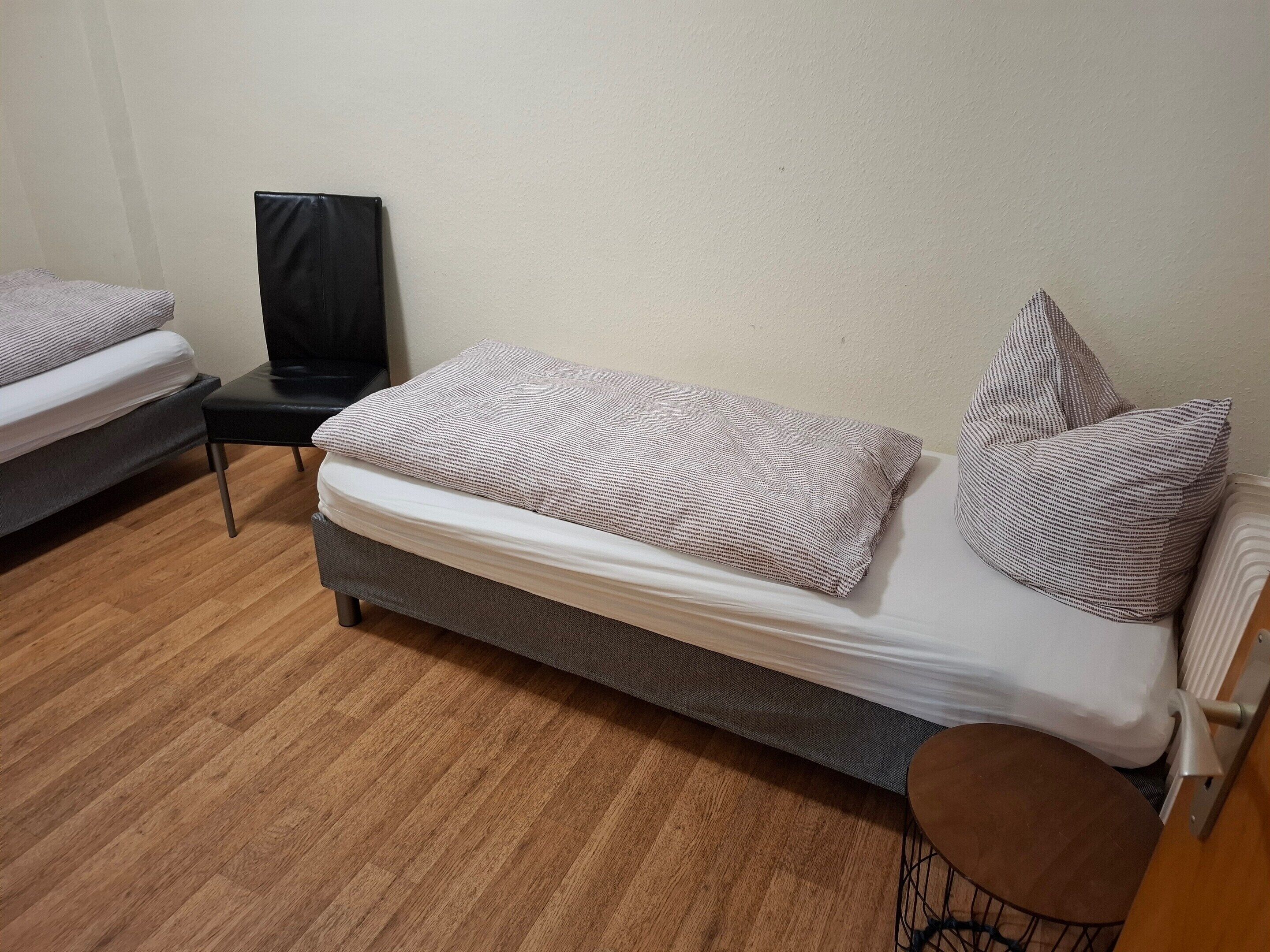 Un lit simple avec une couverture à motifs et des oreillers, une petite chaise noire et un plancher en bois.