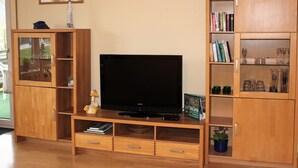 A living room with a flat-screen TV on a wooden stand, wooden cabinets with glass doors, and a small potted plant. - Apartment in Bodenwerder mit Balkon (Bodenwerder)