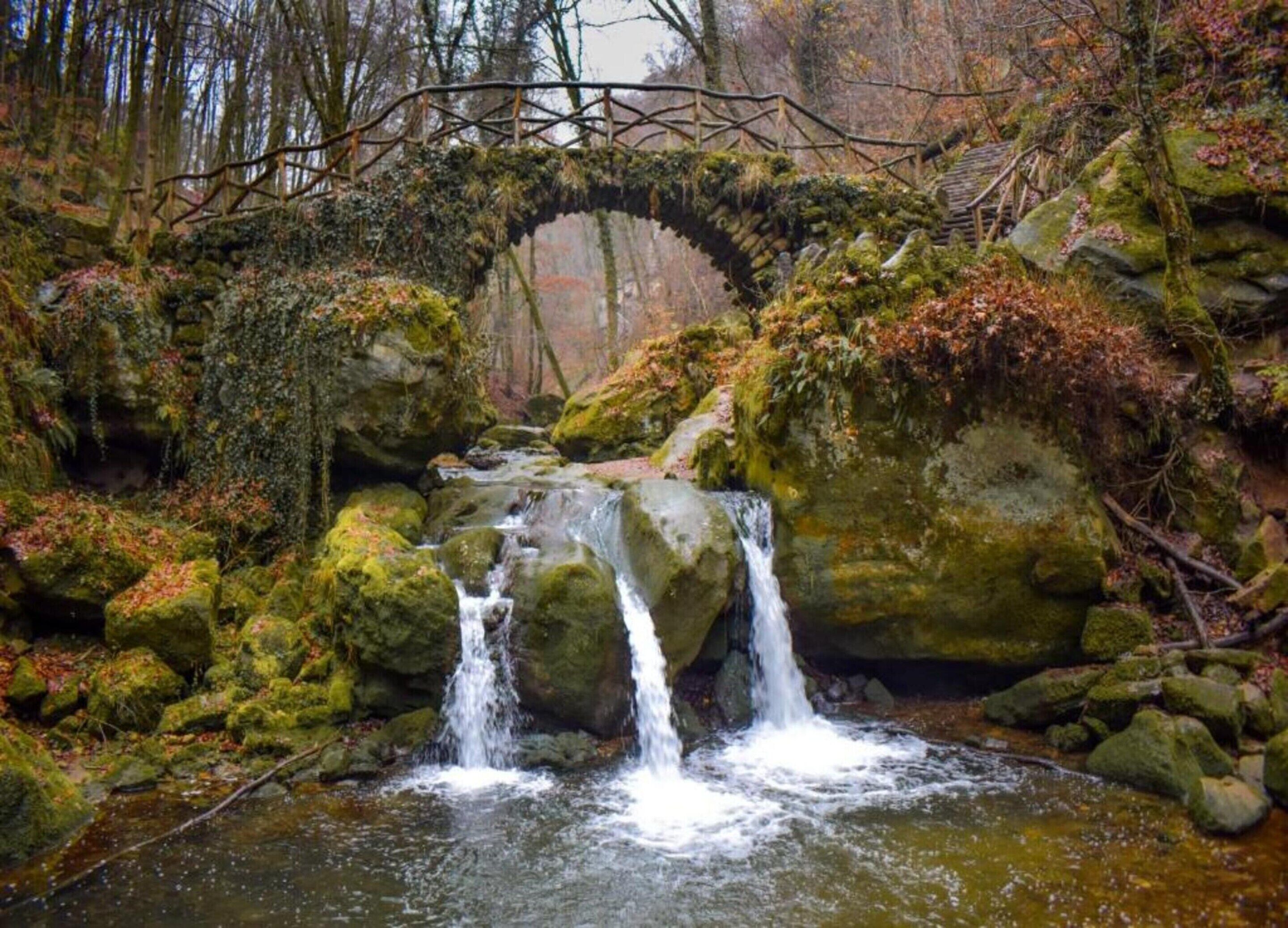 Een landelijke stenen brug over een kleine waterval in een bebost gebied.