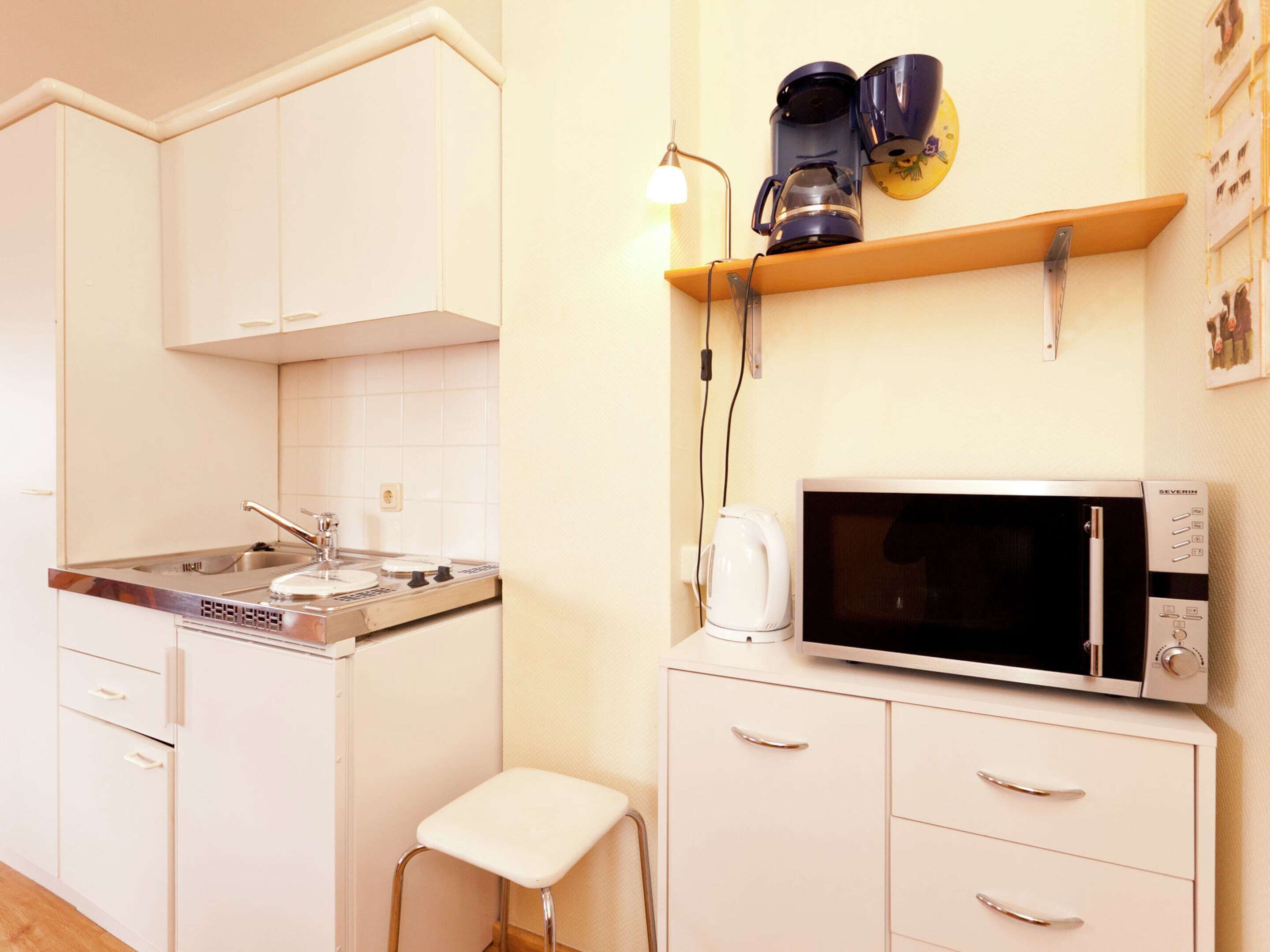 A compact kitchen with white cabinets, a microwave, a coffee maker, and a small stool.