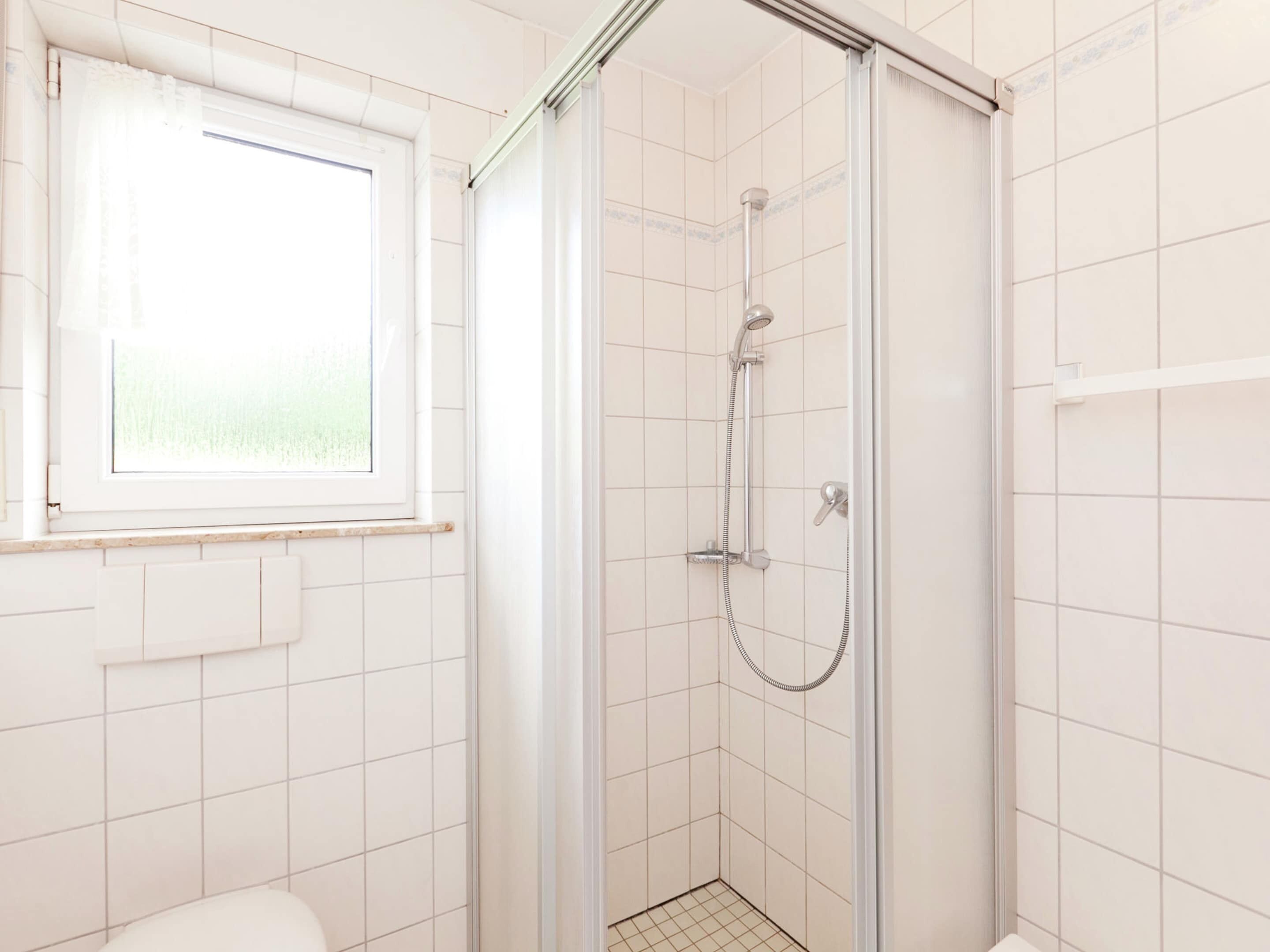 Un baño moderno con ducha de vidrio, paredes de azulejos blancos, una ventana y un inodoro.
