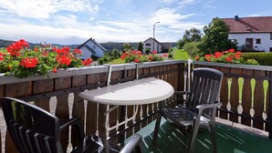A balcony with a round table and chairs, surrounded by a wooden fence, overlooking a village with red flowers and houses. - Modern Apartment in Bolsdorf With Sauna (Hillesheim)