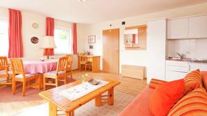 A cozy living room with a wooden table, a sofa, and a dining area with chairs and a table. There is a kitchen area with cabinets and a sink. - Modern Apartment in Bolsdorf With Sauna (Hillesheim)