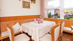 A dining area with a round table, white tablecloth, and wooden chairs. There is a wooden bench along the wall, a shelf with decorative items, and a window with a view of flowers. - Modern Apartment in Bolsdorf With Sauna (Hillesheim)