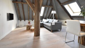 A loft-style room with exposed wooden beams, a grey sofa, a white chair, and a desk. - Apartment Near Groote Keeten Beach (Callantsoog)