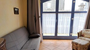 Ein Zimmer mit grauem Sofa, einem gemusterten Ottomanen, einem Holzkabinett und einem großen Fenster mit Vorhängen.