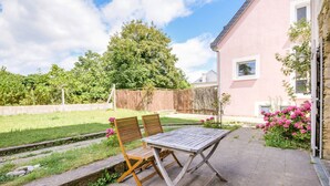 Eine Terrasse im Freien mit Tisch und zwei Stühlen, eine Steinmauer, eine Rasenfläche und ein Gebäude mit Fenstern.