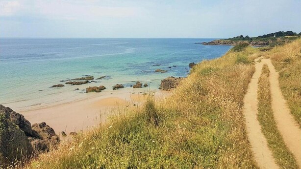 Ein Küstenpfad, der zu einem Strand mit klarem Wasser und weiter Küstenlinie führt.