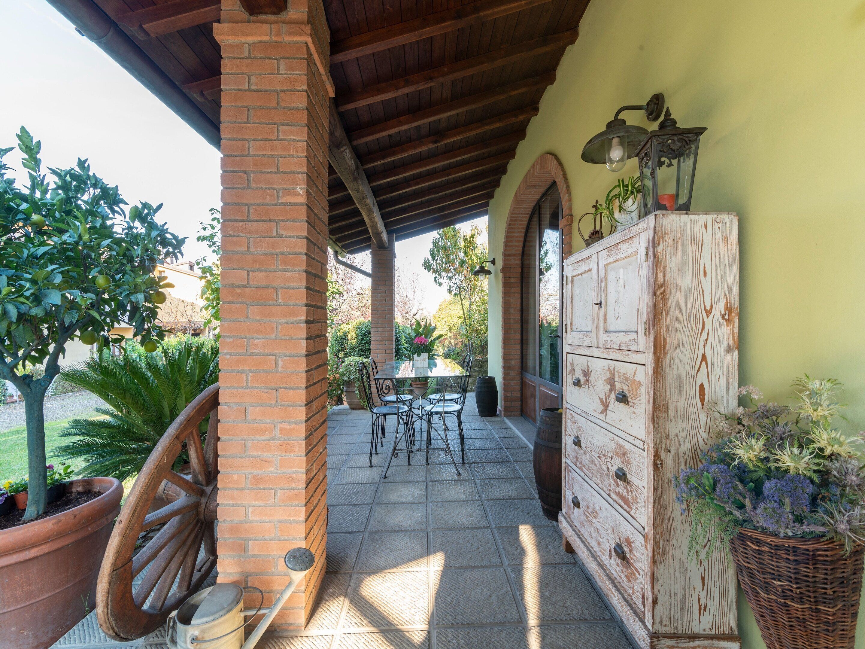 A covered patio area with a brick column, a wooden table with chairs, a vintage dresser, and potted plants.