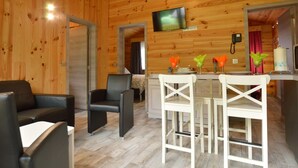 A wooden interior with a TV mounted on the wall, a dining area with white chairs, and a sofa with black cushions. - Chalet Near Durbuy With Private Garden (Ferrieres)