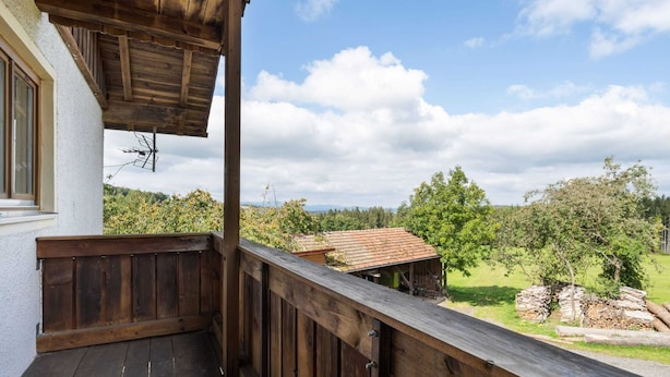 Un balcón de madera con vistas a un paisaje rural, que incluye árboles y un edificio distante.