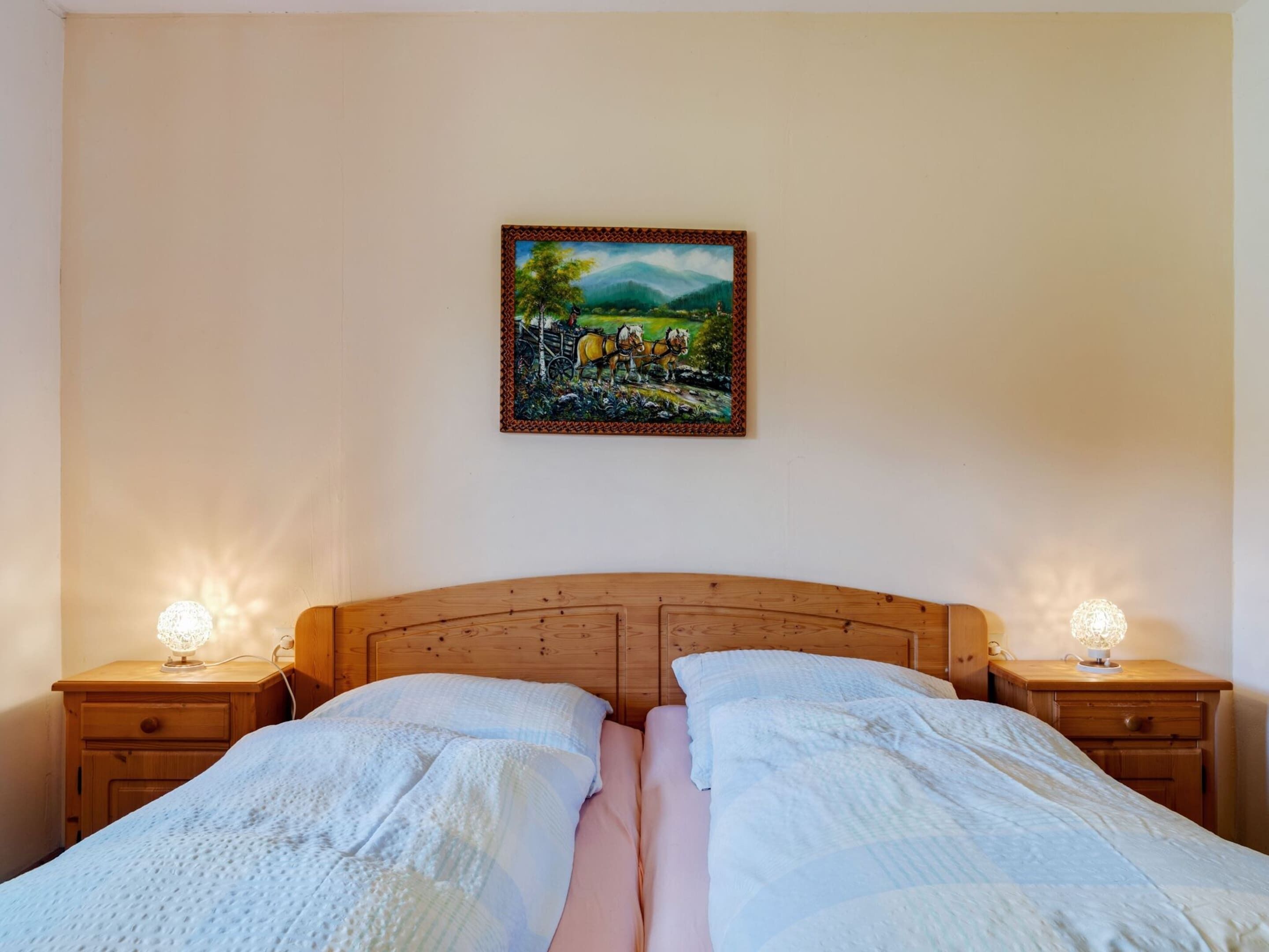 Une chambre à coucher avec une tête de lit en bois, deux tables de chevet avec des lampes, un lit avec une literie blanche et un tableau représentant un paysage rural accroché au mur.