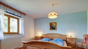 Un dormitorio con cama de madera, dos mesitas de noche con lámparas, un radiador, una ventana con cortinas y un cuadro en la pared.