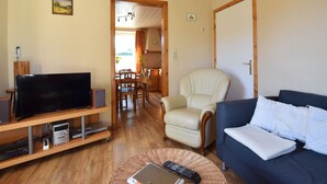 Ein Wohnzimmer mit Fernseher, Sofa, Sessel, Esstisch und einem durchgehenden Türrahmen sichtbaren Küchenbereich.