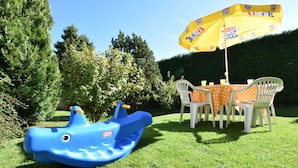 Ein blaues Schlauchboot, ein gelber Sonnenschirm und ein Tisch mit weißen Stühlen in einer grünen Parklandschaft.
