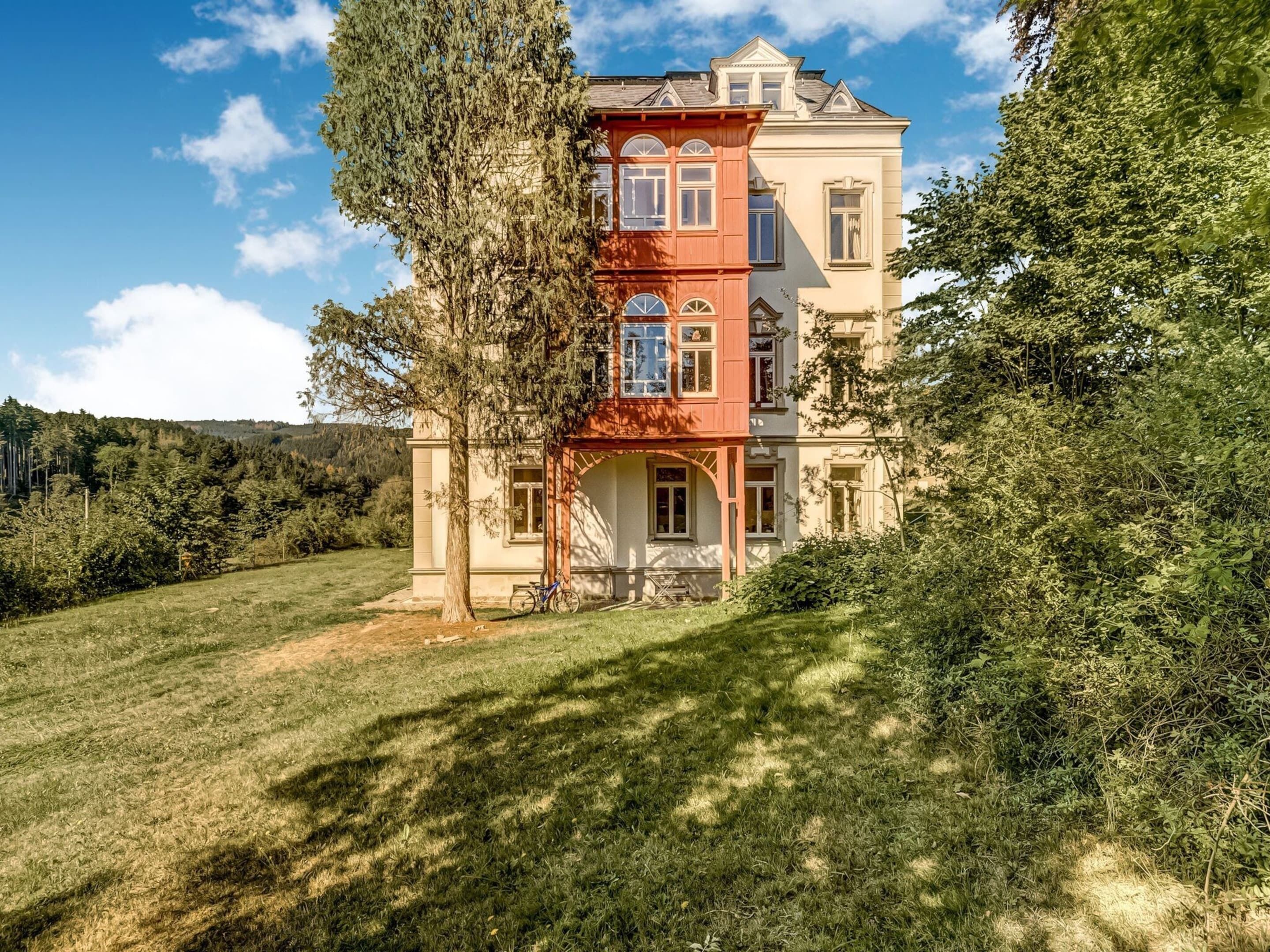 A multi-story building with a prominent red section, surrounded by lush greenery and a clear sky.