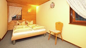 A bedroom with a wooden ceiling, a bed with a headboard, a chair, and a window with lace curtains. - Holiday Home in the Forest (Schmogrow)