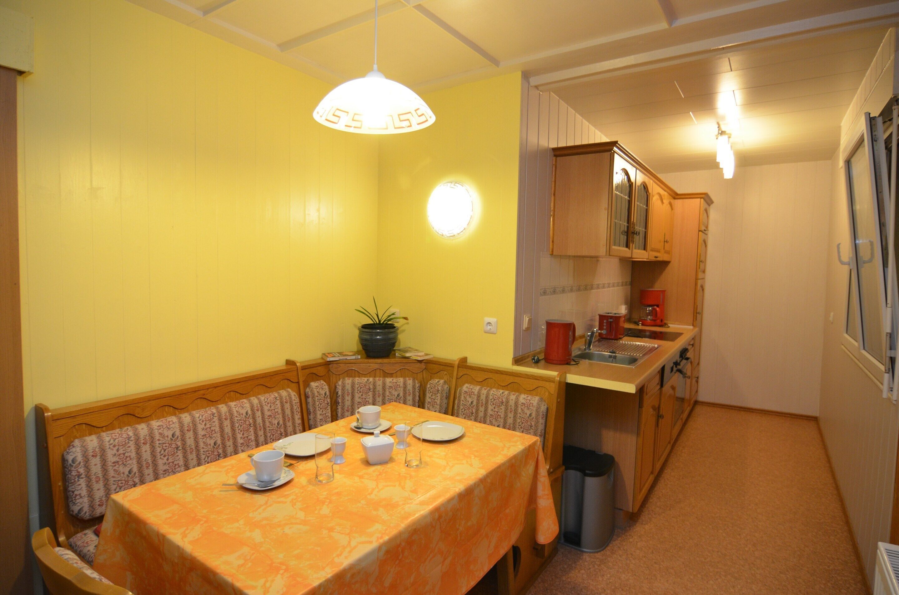 Un comedor con una mesa lista para cuatro personas, una planta en maceta y una cocina al fondo.
