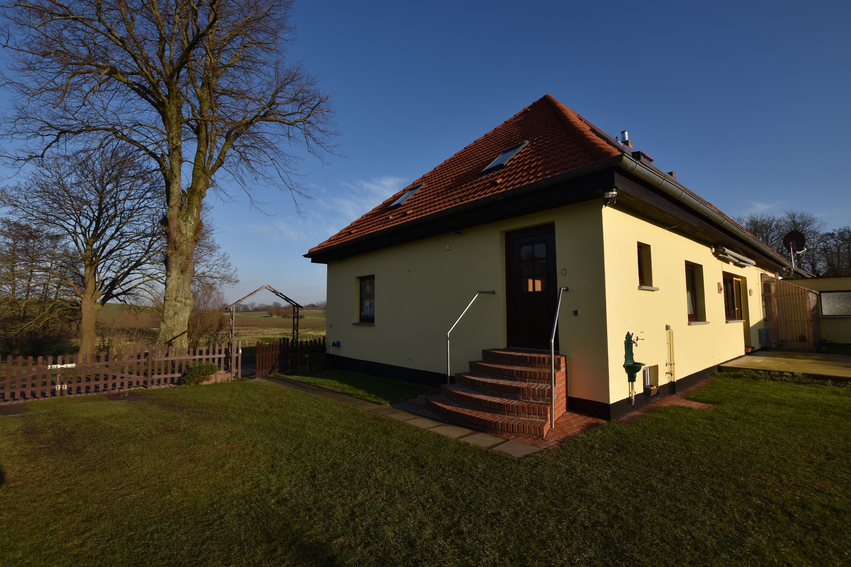 Une maison de plain-pied avec un toit recouvert de tuiles rouges, une porte d’entrée et un petit jardin à l’avant, délimité par une clôture en bois.
