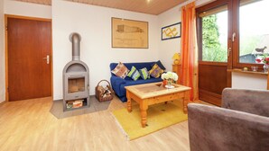 A living room with a wood-burning stove, a blue sofa with patterned cushions, a wooden coffee table, and a window with curtains. - Amazing Apartment in Großalmerode near Cross Country Skiing (Grossalmerode)