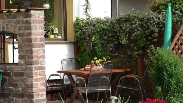 A patio area with a brick column, metal chairs, a wooden table, and potted plants. - Quiet Holiday Home With Terrace (Korbach)